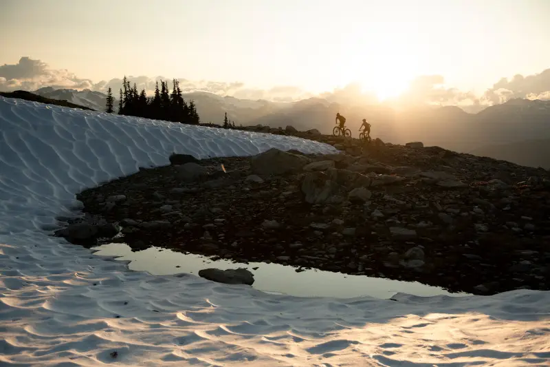 Two cyclists on a mountain trail at sunset with snow and a small pond in the foreground.