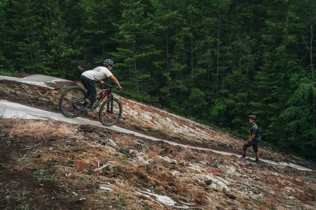 Two individuals mountain biking on a rocky trail surrounded by dense green forest; one rides uphill while the other stands beside his bike observing.
