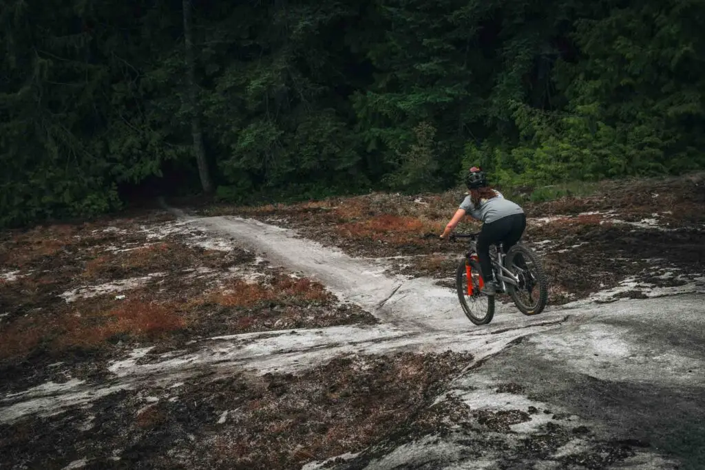 A cyclist wearing a helmet rides a mountain bike on a dirt path through a forested area.